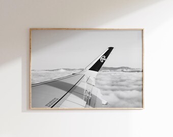 May include: Monochrome framed print showing an airplane wing and tail fin. The tail fin features a logo. The wing is angled upwards, with a sea of clouds and mountains in the background.