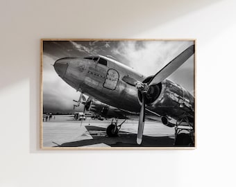 Op de afbeelding: Zwart-witfoto van een vintage vliegtuig op een landingsbaan. Het vliegtuig heeft grote propellers en het woord "AIR" is zichtbaar aan de zijkant. De afbeelding is ingelijst in een houten lijst.