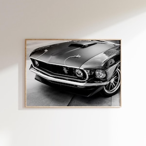 May include: Black and white framed print of a classic black muscle car. The car features a hood scoop, chrome accents, and a prominent front grille. The image is a close-up of the front of the vehicle, showcasing its headlights and wheels.