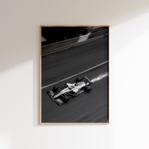 May include: Monochrome framed print of a Formula 1 race car speeding along a track. The car is white with black wheels and accents, captured mid-turn with motion blur. Sparks trail from the rear, emphasizing speed and action. The print is in a wooden frame.