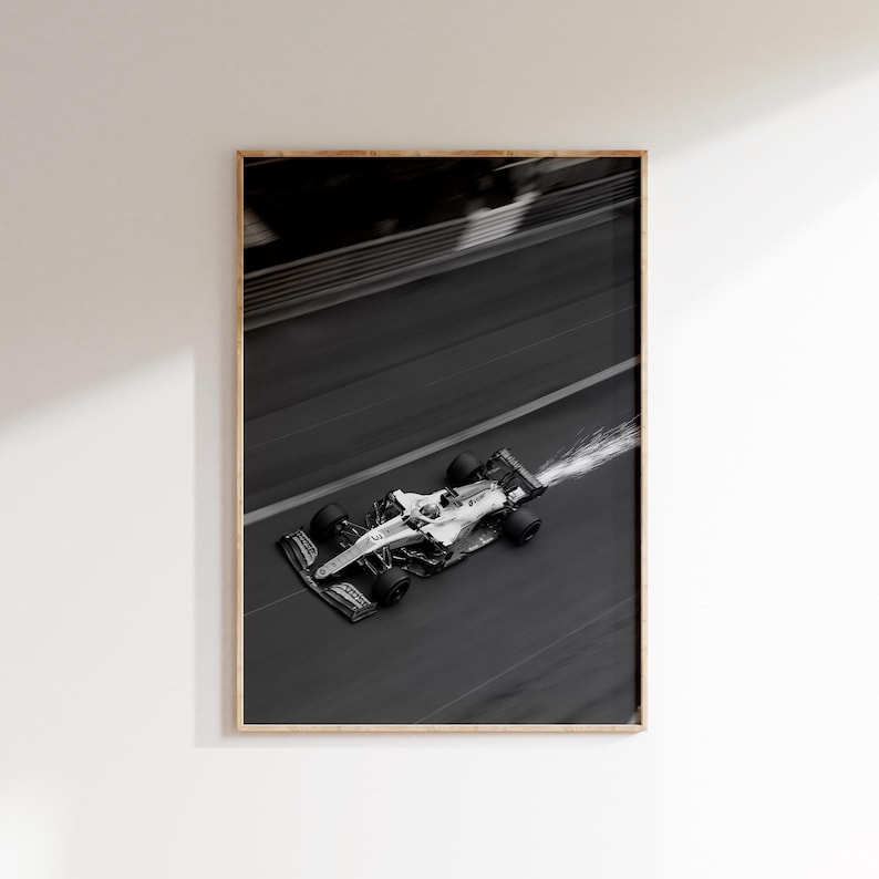 May include: Monochrome framed print of a Formula 1 race car speeding along a track. The car is white with black wheels and accents, captured mid-turn with motion blur. Sparks trail from the rear, emphasizing speed and action. The print is in a wooden frame.
