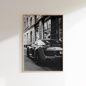 May include: Black and white framed print of a sleek sports car parked on a city street. The car's design is emphasized by the monochrome tones, with a focus on its aerodynamic features and rear profile. The background shows a classic building facade.