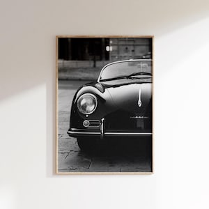 May include: Framed monochrome print of a vintage car. The image focuses on the front of the car, showcasing a round headlight, chrome bumper, and hood ornament. The car's glossy black finish is highlighted in this close-up shot.
