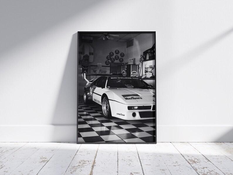 May include: Black and white framed print of a race car in a garage. The car is white with black accents and the Marlboro logo. The garage has a checkered floor and Pepsi signage.