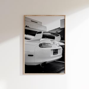 Puede incluir: Fotografía en blanco y negro de un coche deportivo Toyota Supra blanco. Se ve la parte trasera del coche, con el alerón, las luces traseras y el tubo de escape. La matrícula muestra "122".