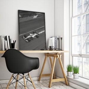 May include: A framed black and white photograph of a Formula 1 race car speeding along a track. The image is displayed on a wooden desk, alongside books, a black chair, and decorative items. The scene is illuminated by natural light from a nearby window.