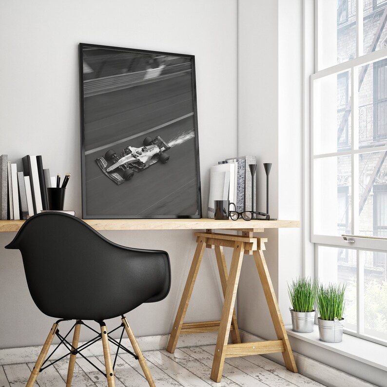 May include: A framed black and white photograph of a Formula 1 race car speeding along a track. The image is displayed on a wooden desk, alongside books, a black chair, and decorative items. The scene is illuminated by natural light from a nearby window.