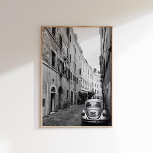 Puede incluir: Impresión enmarcada en blanco y negro de una calle europea estrecha. Un coche antiguo está estacionado en la calle adoquinada, mirando al espectador. Los edificios bordean ambos lados de la calle, con ventanas y balcones. La impresión está en un marco de madera.