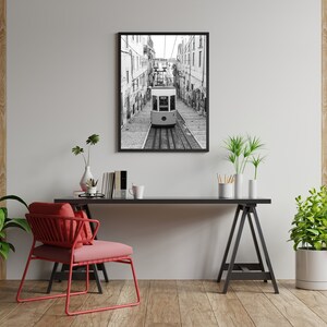 May include: Black and white framed print of a funicular railway car traveling down a cobblestone street. The image is displayed above a black desk with a red chair, plants, and decorative items. The room has wooden floors and a light gray wall.