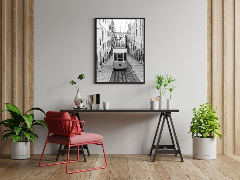 May include: Black and white framed print of a funicular railway car traveling down a cobblestone street. The image is displayed above a black desk with a red chair, plants, and decorative items. The room has wooden floors and a light gray wall.