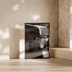 May include: Monochrome framed print depicting a Formula 1 car in action on a race track. The print is propped against a neutral wall, creating a minimalist aesthetic. The image captures the dynamic movement of the car, highlighting motorsport.