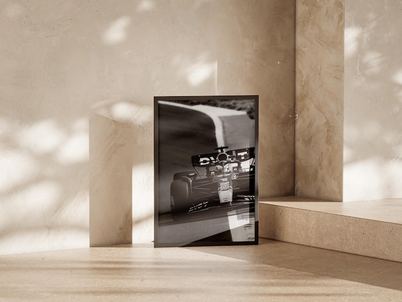 May include: Monochrome framed print depicting a Formula 1 car in action on a race track. The print is propped against a neutral wall, creating a minimalist aesthetic. The image captures the dynamic movement of the car, highlighting motorsport.