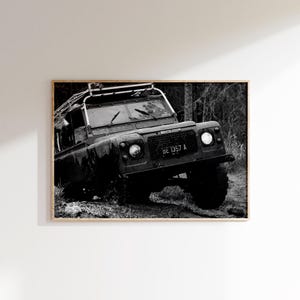 Puede incluir: Impresión enmarcada en blanco y negro de un Land Rover todoterreno vintage. El vehículo oscuro tiene baca y la matrícula es BE 1357 A. La imagen está ambientada en un entorno boscoso y embarrado.