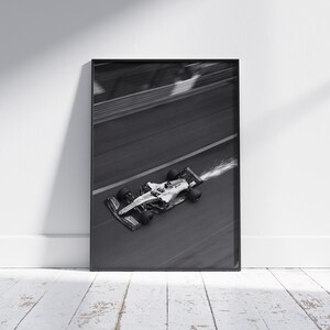 May include: Black and white framed print of a Formula 1 race car speeding along a track. The car is in motion, with motion blur and a trail of vapor. The image is set against a white wall and a wooden floor.