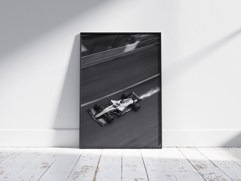 May include: Black and white framed print of a Formula 1 race car speeding along a track. The car is in motion, with motion blur and a trail of vapor. The image is set against a white wall and a wooden floor.