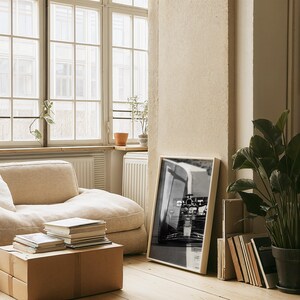 May include: A light-filled living space featuring a beige beanbag chair, a framed monochrome photograph, and a large window. A stack of books rests on a cardboard box, and a potted plant adds a touch of greenery to the room.