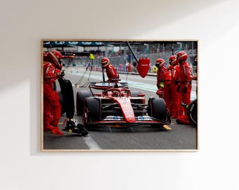 May include: A framed print of a Formula 1 race car in a pit stop. The car is red with black tires and accents. Crew members in red uniforms and helmets surround the car, preparing for the pit stop. The background shows the race track and other team members.