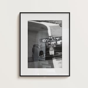 May include: Monochrome framed print of a Formula 1 car racing on a track. The car is captured mid-turn, with the track's white lines in view. The print is presented in a black frame.