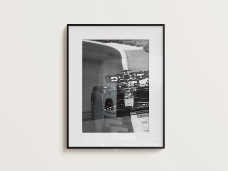 May include: Monochrome framed print of a Formula 1 car racing on a track. The car is captured mid-turn, with the track's white lines in view. The print is presented in a black frame.
