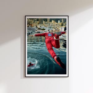 Può includere: Una fotografia incorniciata mostra una persona con una tuta da corsa rossa con logo HP che salta in acqua. La persona è a mezz'aria con le braccia tese, mentre una seconda persona in tuta rossa è visibile nell'acqua. Lo sfondo mostra un porto.