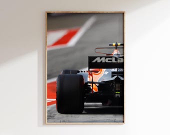 May include: A framed print featuring a close-up of a Formula 1 race car. The car is black, orange, and silver, with the McLaren logo visible. The image captures the car's rear tire and part of the body, set against a blurred track with red and white markings.
