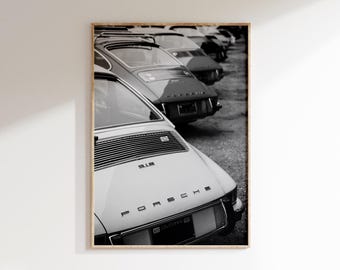 May include: Black and white photograph of a row of classic Porsche 911 cars. The cars are parked in a line, with the rear of the vehicles facing the viewer. The word "PORSCHE" is visible on the back of one car.