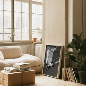 May include: A framed black and white photograph of a race car on a track. The photograph is leaning against a wall in a room with a beige sofa, a cardboard box, and a stack of books. A large potted plant is in the corner.