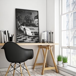 May include: Black and white framed print of a vintage race car in a garage setting. The car is white with sponsor logos, parked on a checkered floor. The print is displayed on a wooden desk with books, a black chair, and a window.