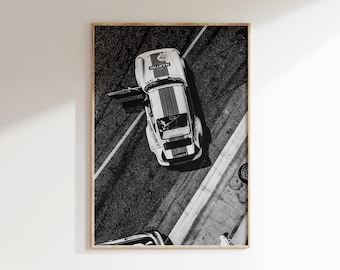 May include: Black and white framed print of a vintage race car with the door open, viewed from above. The car has a white body with red and blue stripes and the word "MARTINI" on the hood. The car is on a racetrack.