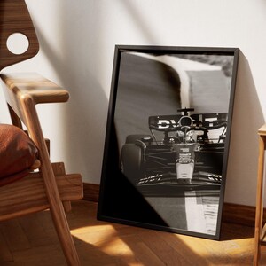 May include: Monochrome framed print of a Formula 1 racing car on a track. The car is captured in motion, with the front of the vehicle in focus. The print is propped against a white wall, beside a wooden chair.
