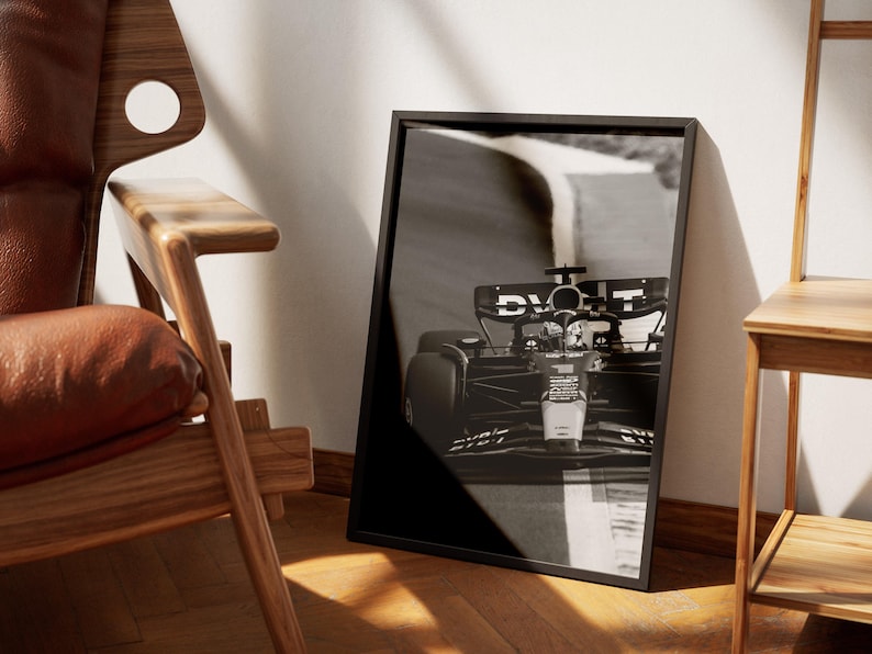 May include: Monochrome framed print of a Formula 1 racing car on a track. The car is captured in motion, with the front of the vehicle in focus. The print is propped against a white wall, beside a wooden chair.