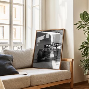 May include: Monochrome framed print of a Formula 1 racing car on a track. The wooden-framed picture leans against a beige sofa. The image captures the speed and motion of the car.