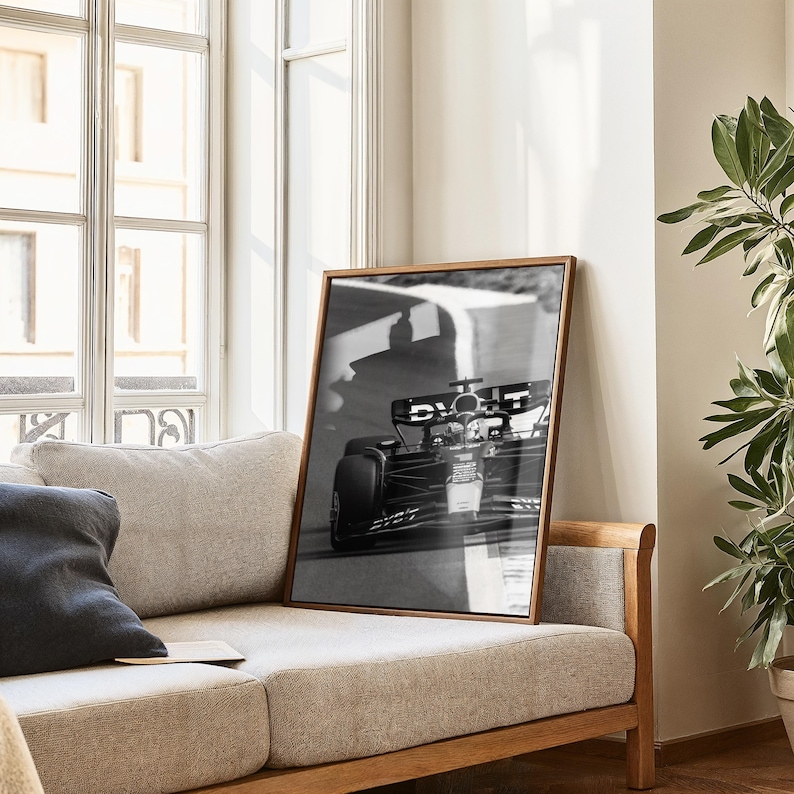 May include: Monochrome framed print of a Formula 1 racing car on a track. The wooden-framed picture leans against a beige sofa. The image captures the speed and motion of the car.
