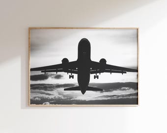 May include: A monochrome framed print featuring the silhouette of an aeroplane against a cloudy sky. The aircraft is depicted in flight, with its wings and engines clearly defined. The artwork has a minimalist style.