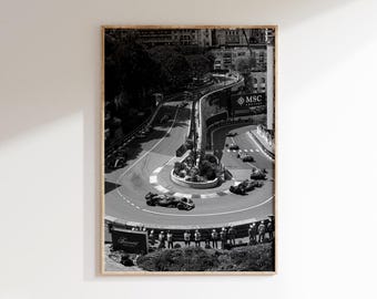 May include: Black and white framed print of a Formula 1 race. The image captures a race track with multiple race cars navigating a sharp turn. Spectators line the track, and buildings are visible in the background.