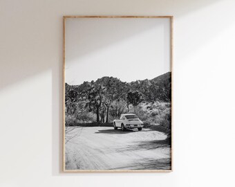 May include: Black and white framed print featuring a vintage white sports car parked on a dirt road. The car is set against a backdrop of desert vegetation and a mountain range under a bright sky. The print is in a wooden frame.