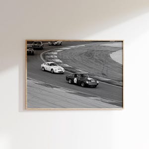 May include: Black and white framed print of vintage race cars on a track. The image shows several classic cars in a race, with one car in the foreground. The cars are racing around a curved track.