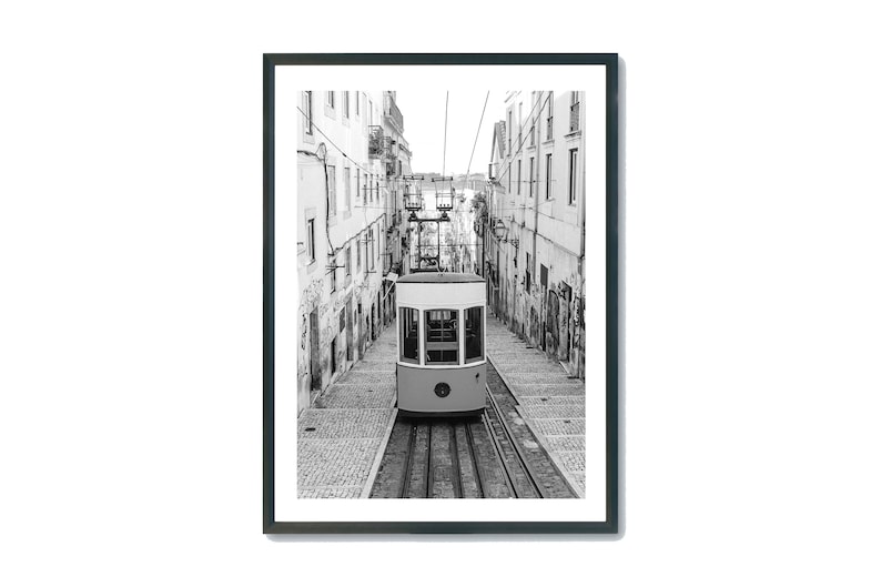 May include: Black and white framed print of a funicular ascending a cobblestone street. The funicular is cream-colored with a white roof and large windows. Buildings line the street on both sides.