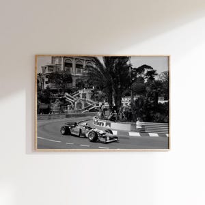 May include: Black and white framed photograph of a Formula 1 race car speeding around a track. The car is branded with the Marlboro logo. The background features a building and lush greenery.