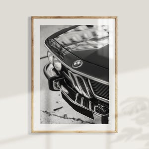 May include: Black and white framed print featuring a close-up of a classic car's front end. The image highlights the car's hood, grille, and emblem. The glossy black paint reflects the surroundings, creating a striking contrast.