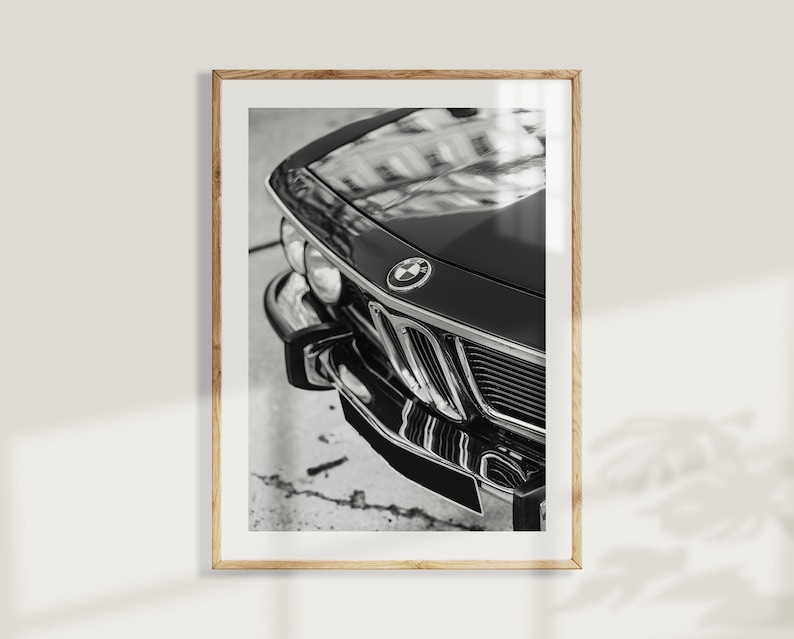 May include: Black and white framed print featuring a close-up of a classic car's front end. The image highlights the car's hood, grille, and emblem. The glossy black paint reflects the surroundings, creating a striking contrast.