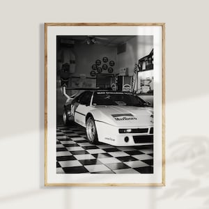 May include: Black and white framed print of a vintage race car. The car is white with black accents and sponsor logos, including "Marlboro." The car is parked in a garage with a checkered floor.