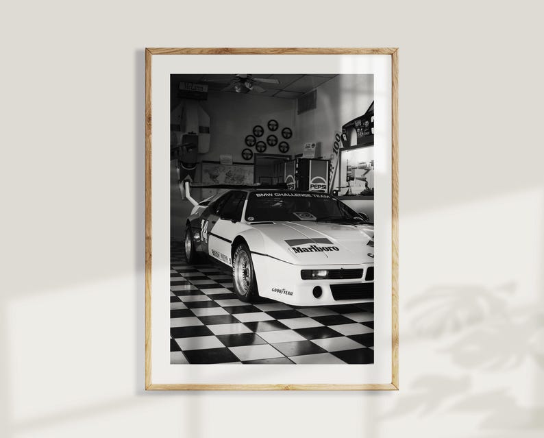 May include: Black and white framed print of a vintage race car. The car is white with black accents and sponsor logos, including "Marlboro." The car is parked in a garage with a checkered floor.
