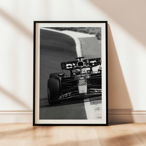 May include: Monochrome framed print depicting a Formula 1 car navigating a bend on a race track. The image captures the car's details, including tyres and cockpit. The print is displayed against a white wall and a wooden floor.