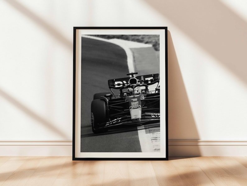 May include: Monochrome framed print depicting a Formula 1 car navigating a bend on a race track. The image captures the car's details, including tyres and cockpit. The print is displayed against a white wall and a wooden floor.