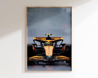 May include: Framed print of a Formula 1 race car in motion. The car is orange and black, with a driver visible inside. The background is a blurred, overcast sky, suggesting a rainy race day.