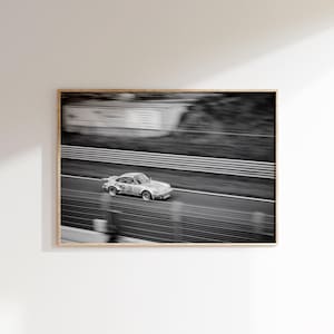 以下が含まれることがあります： 白黒フレームプリント。銀色のレーシングカーがトラックを走行しています。車のスピードを捉え、トラックと白とグレーのバリアがぼやけています。