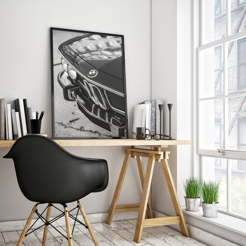 May include: Black and white framed print of a classic car's front end, featuring the BMW logo. The print is displayed on a wooden desk with a black chair, books, and decorative items. The scene is illuminated by natural light from a nearby window.