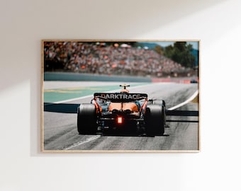 May include: Framed print of a Formula 1 race car on a track. The car is orange and black, with the word "DARKTRACE" visible. The background shows a crowd of spectators and green trees.