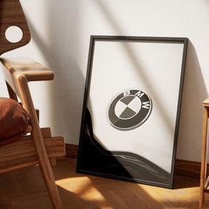 May include: Framed print featuring the BMW logo. The logo is a black and white circular emblem with the letters BMW. The print is in a black frame, leaning against a white wall. A wooden chair and side table are in the background.